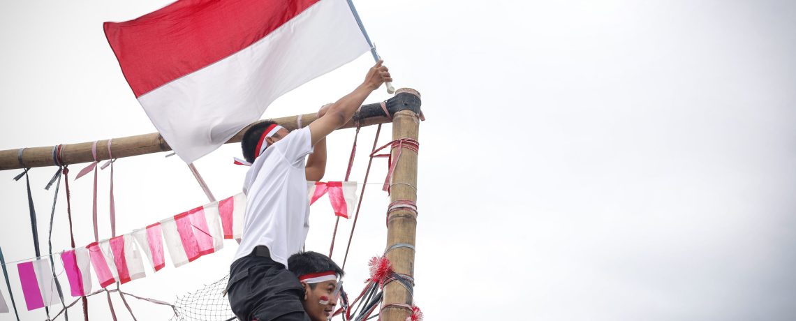 Two Young Indonesian Man Hoisting The Indonesian Flag On Tall Bamboo Pole During Traditional Festivities