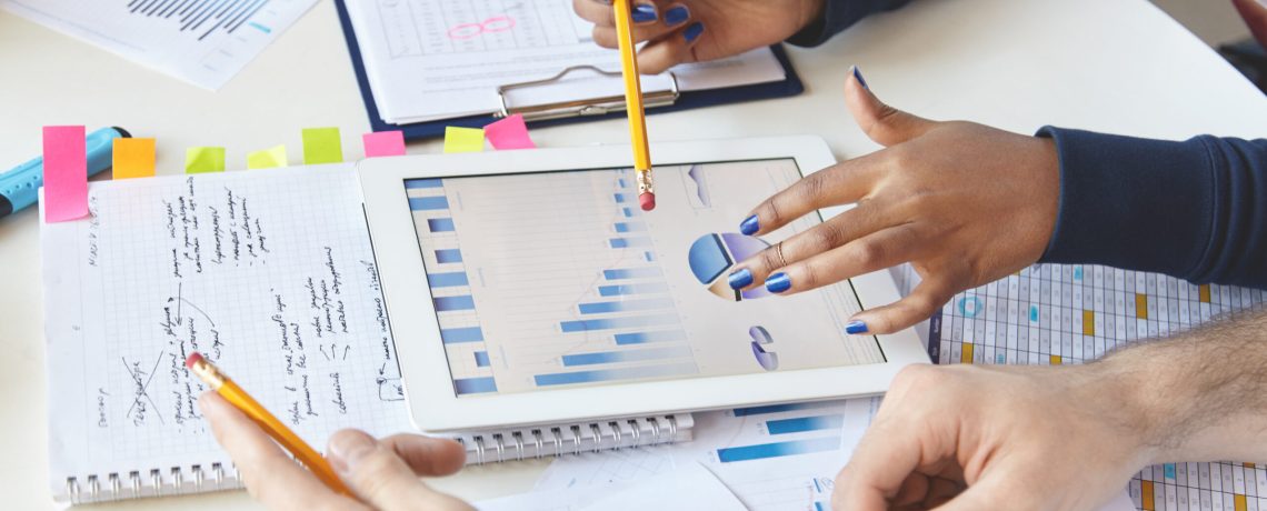 Colleagues working together on financial report using modern gadget. African girl with pencil presenting information on laptop screen, studying company's growth, showing diagram to her partners